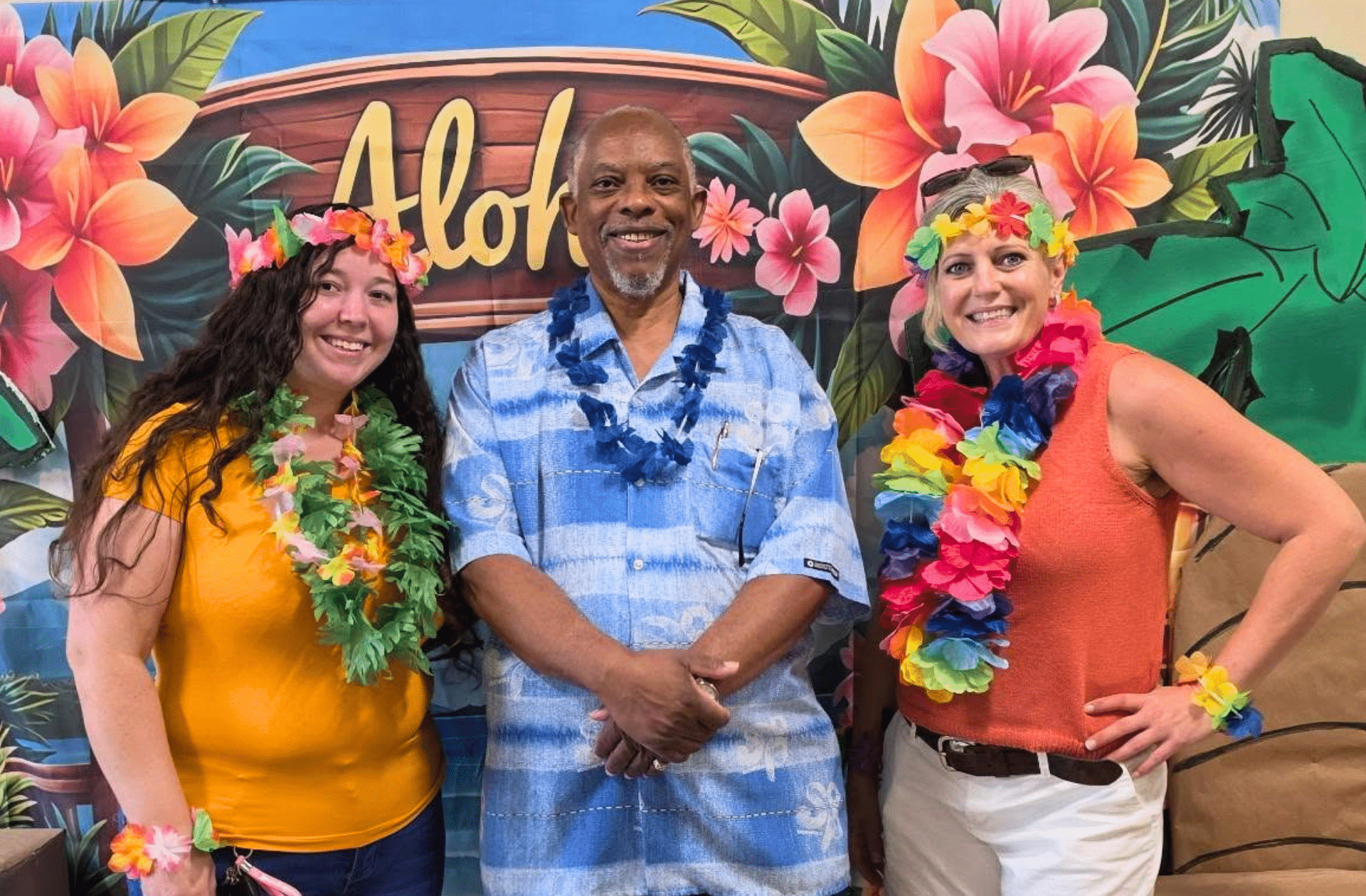 Members of the Family Connection Camden Leadership Team standing in front of a colorful tropical backdrop with large flowers, leaves, and the word ‘Aloha.’ All three are wearing bright, multicolored leis and tropical-themed clothing.